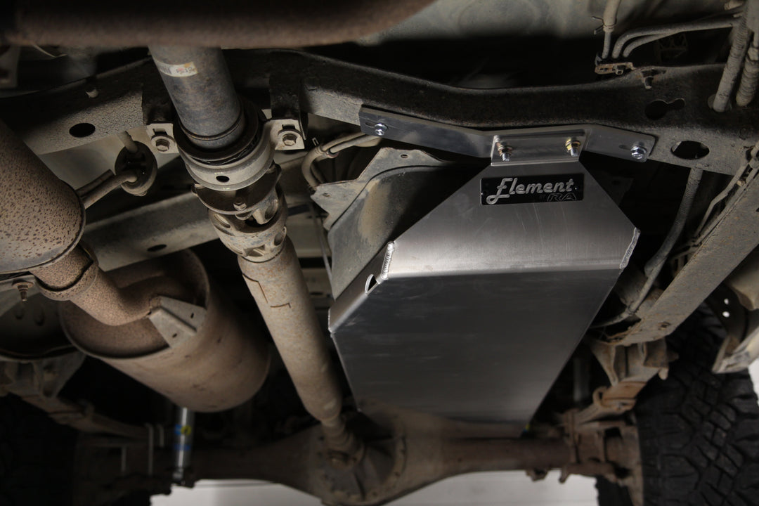 00-04 TOYOTA TUNDRA ALUMINUM GAS TANK SKID PLATE The image shows the underside of a vehicle with a focus on an installed RA Motorsports gas tank skid plate. The skid plate is metallic with a shiny surface and is securely attached to the undercarriage, providing protection to the gas tank area. Visible parts include exhaust pipes and other mechanical components, which appear used and dirty. The brand “Element” is displayed on the skid plate.
